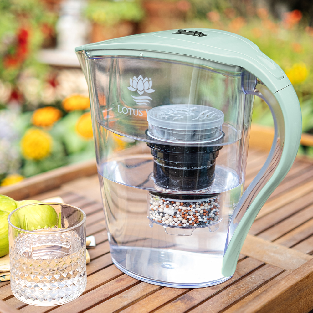 Eine zartgrüne Wasserfilterkanne von Lotus Vita steht auf einem Gartentisch, gefüllt mit gefiltertem Wasser, daneben ein Trinkglas.