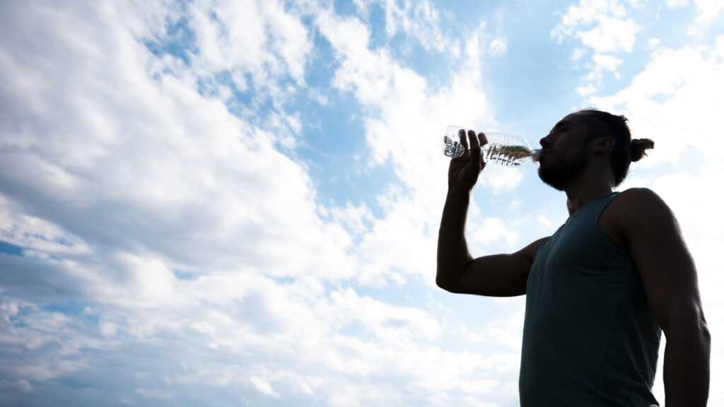 Ein sportlicher Mann erfrischt sich nach dem Training und trinkt reines Wasser aus einer Flasche, im Hintergrund ein blauer Himmel mit Wolken.