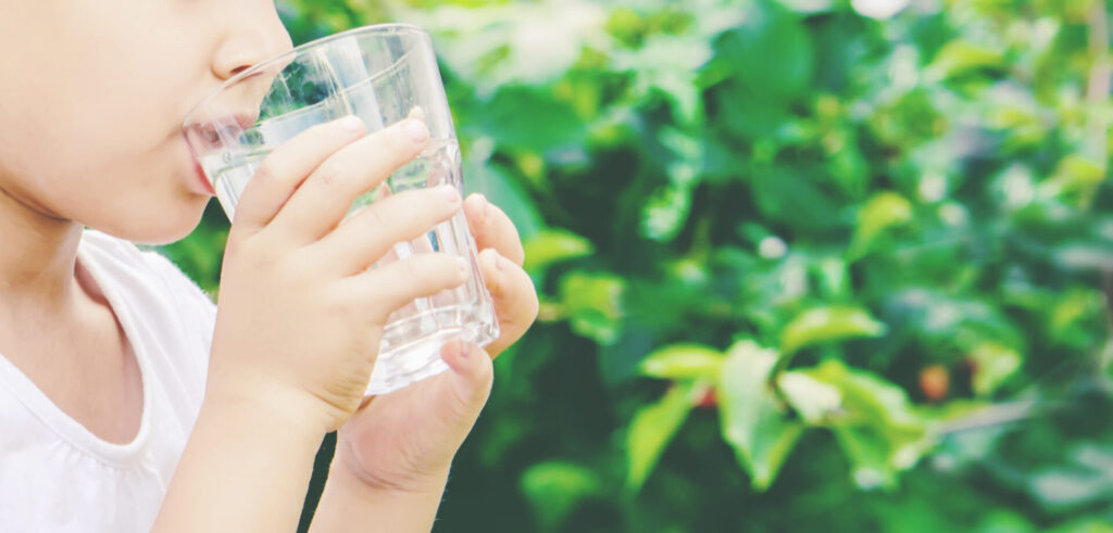Ein Kind in einem weißen T-Shirt trinkt klares Wasser aus einem Glas, vor einem grünen, natürlichen Hintergrund.