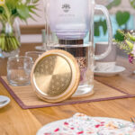Eine stilvolle Glas-Wasserfilterkanne von Lotus Vita steht auf einem gedeckten Holztisch, davor liegt der goldene Bambusdeckel mit einer Gravur der Lebensblume.