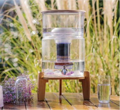 Ein Glas-Wasserfilter mit Bambussockel und -deckel steht auf einem Holztisch im Freien, daneben ein Glas mit gefiltertem Wasser.