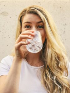 Eine junge Frau mit blonden Haaren und einem weissen T-Shirt blickt direkt in die Kamera, während sie ein Glas mit Wasser und Eiswürfeln trinkt.