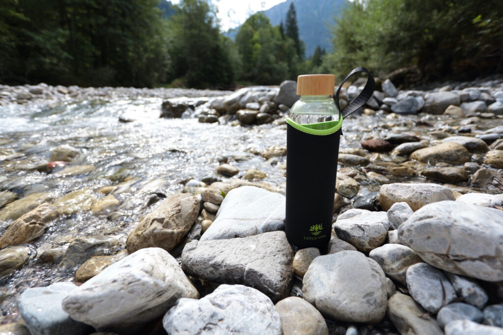 Eine Glas-Trinkflasche mit schwarzer Schutzhülle und Holzdeckel steht auf den Steinen eines klaren Gebirgsbachs im Allgäu.