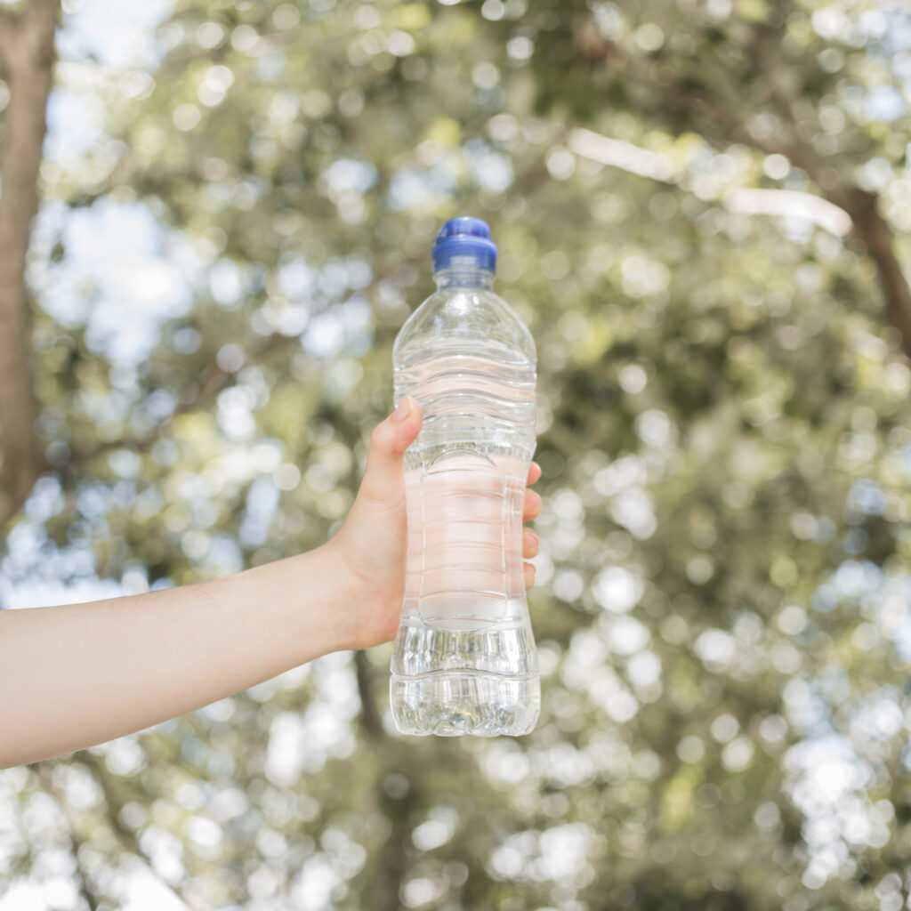 Eine Hand hält eine durchsichtige Plastik-Wasserflasche vor einem unscharfen, grünen Hintergrund im Freien.