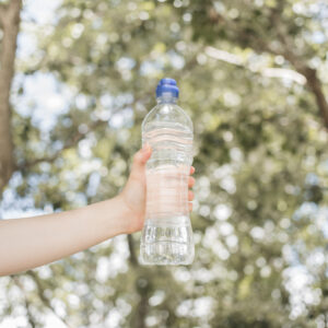 Eine Hand hält eine durchsichtige Plastik-Wasserflasche vor einem unscharfen, grünen Hintergrund im Freien.