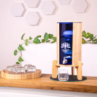 Ein Glas-Wasserfilter mit Bambusgehäuse und -ständer steht auf einer Holztheke, daneben ein Tablett mit Gläsern und eine grüne Pflanze im Hintergrund.