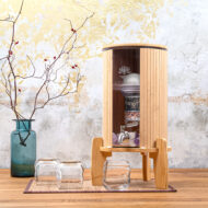 Ein Wasserfilter aus Glas und Bambus von Lotus Vita steht auf einem Holztisch, daneben zwei Gläser und eine Vase mit Hagebuttenzweigen.