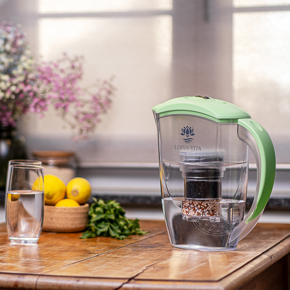 Eine Wasserfilter-Kanne mit hellgrünem Deckel steht auf einem Holztisch neben einem Glas Wasser, frischen Zitronen und Minze in einer hellen Küche.